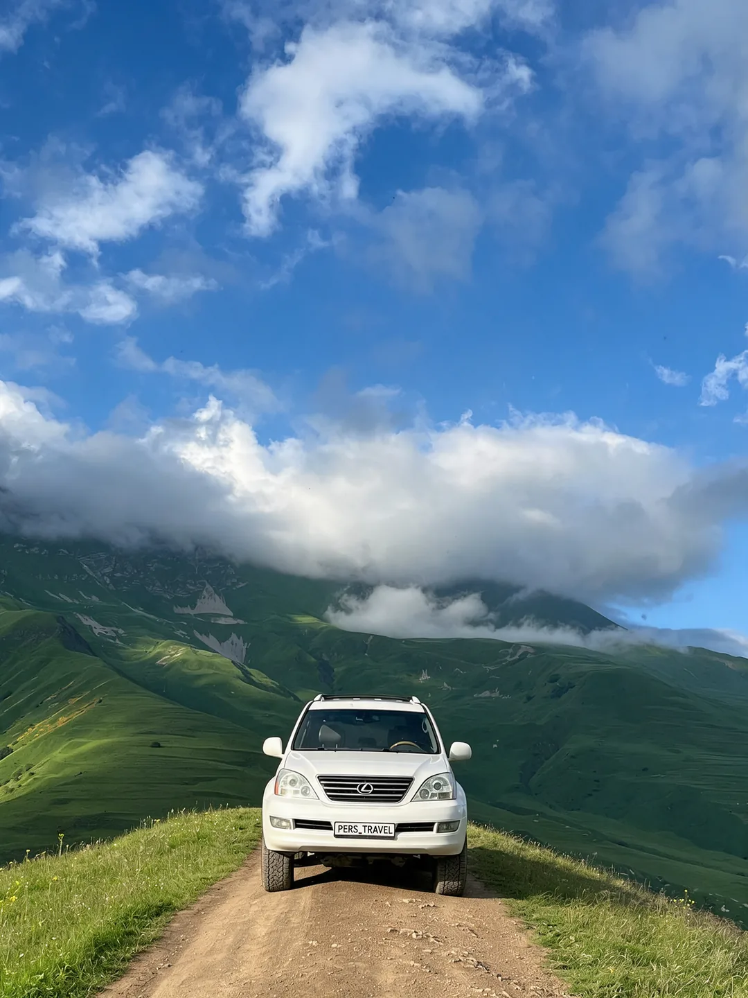 Белый Lexus Аслана с номером PERS_TRAVEL на грунтовой дороге по горному хребту, облака над вершиной